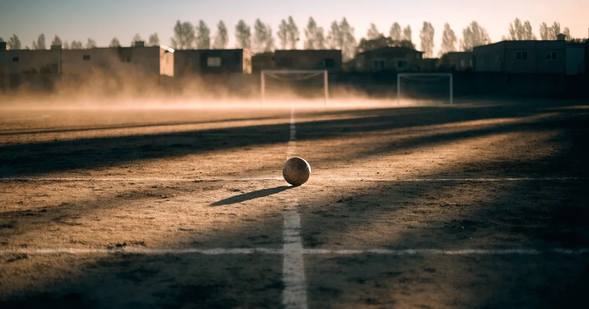 Ein sandiger Fußballplatz am Rand einer Stadt in warmem Abendlicht, Staub in der Luft und zwei Tore in der Ferne.