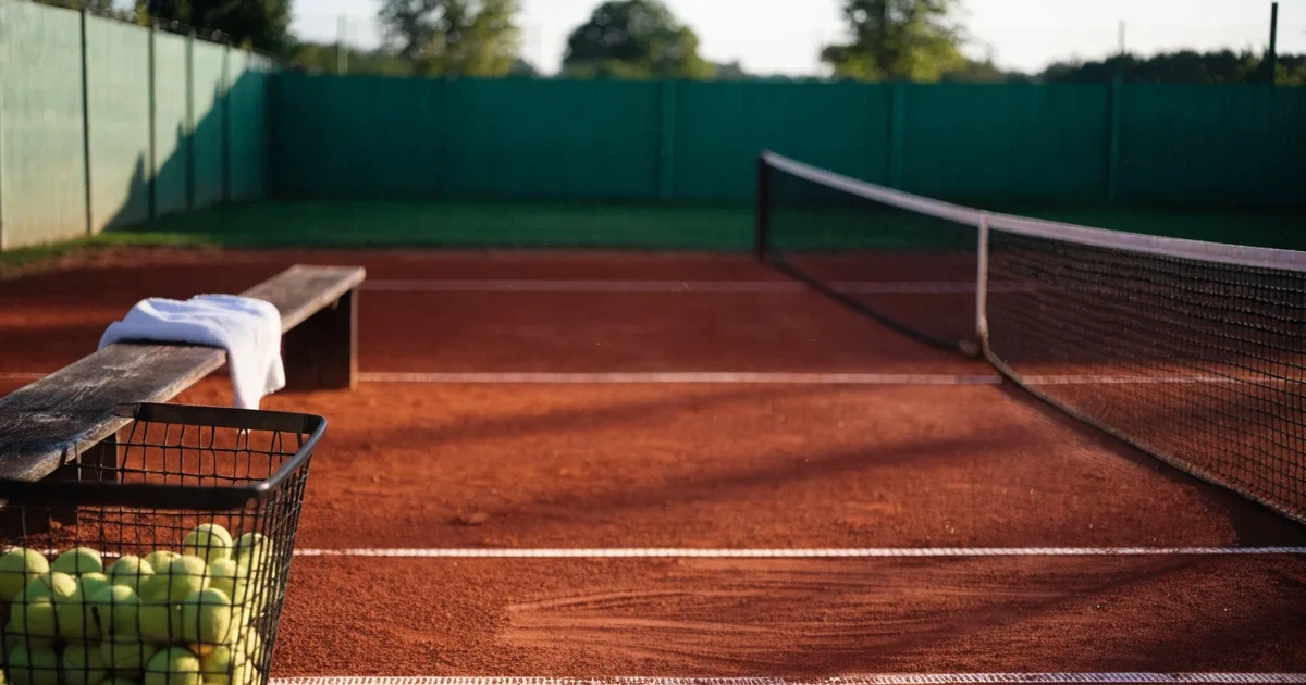 Ein Sandplatz mit frisch gezogenen Linien in der Nachmittagssonne, Netz und Ballkorb am Rand, keine Menschen.