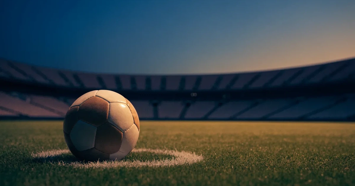 Ein Fußball liegt auf dem Anstoßpunkt eines leeren Rasenplatzes, im Hintergrund eine große Stadiontribüne im Abendlicht.