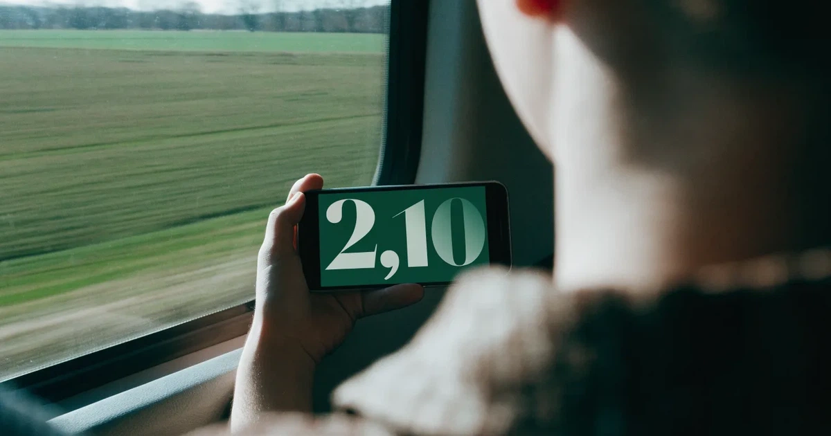 Eine Hand hält ein Smartphone mit dunklem Display in einem Zug, am Fenster ziehen verschwommen Felder vorbei.