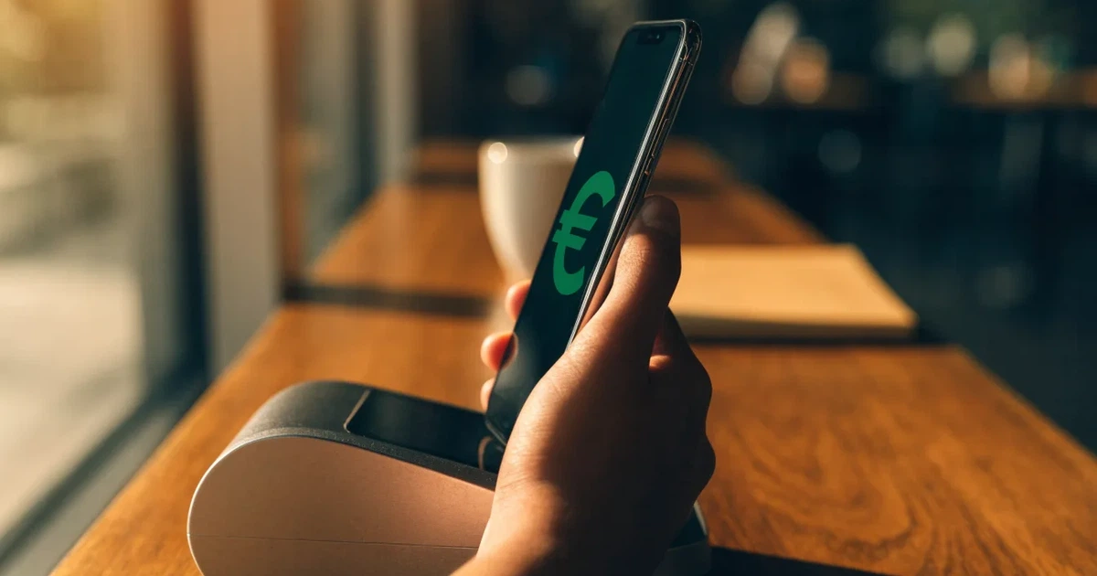 Eine Hand hält ein Smartphone dicht an ein Kartenlesegerät ohne erkennbare Beschriftung in einem Café.