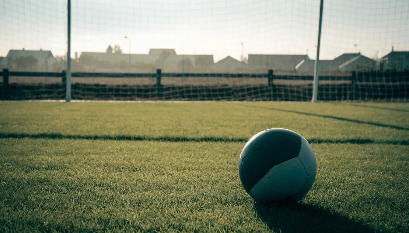Ein Fußball auf dem Rasen eines leeren Sportplatzes am Rand einer Kleinstadt im Morgenlicht.