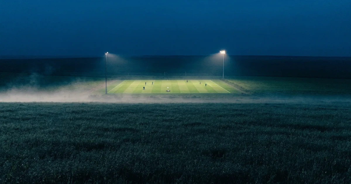 Ein Stadion unter Flutlicht, aus großer Entfernung über eine dunkle Wiese fotografiert, am Himmel zieht Nebel auf.