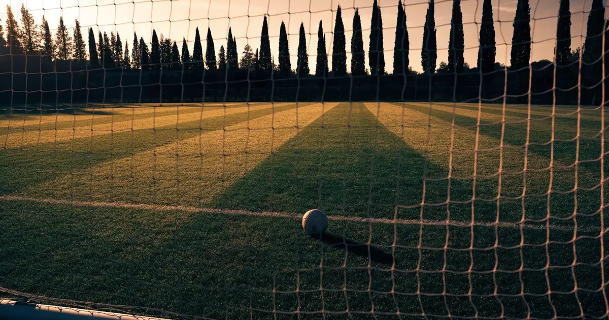 Ein Fußballtor mit Netz auf einem leeren Platz im Abendlicht, der Rasen frisch gemäht, im Hintergrund Zypressen.