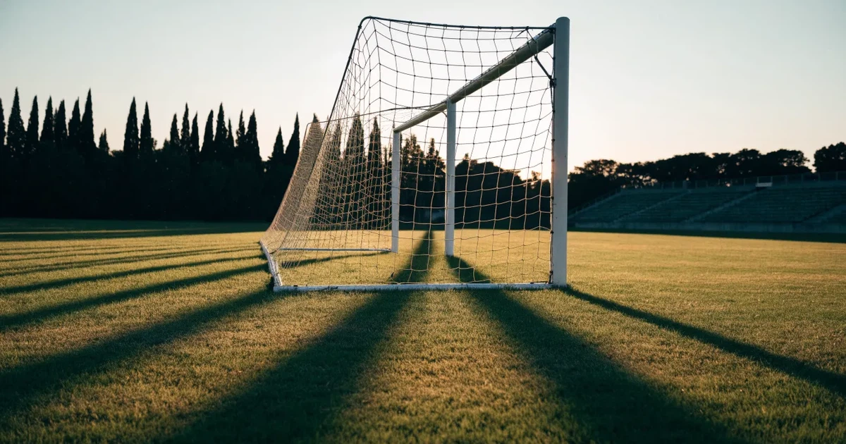 Ein Fußballtor mit Netz auf einem leeren Platz im Abendlicht, der Rasen frisch gemäht, im Hintergrund Zypressen.