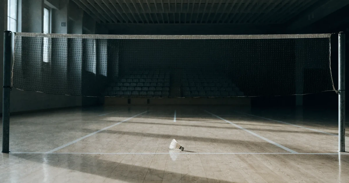 Eine leere Badmintonhalle mit gespanntem Netz und hellen Hallenlichtern, ein Federball liegt auf dem Hallenboden.