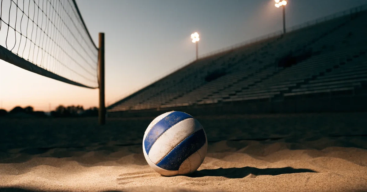 Ein Volleyball liegt auf dem Sand eines Beachvolleyball-Feldes neben dem gespannten Netz, im Hintergrund eine leere Tribüne bei Abendlicht.