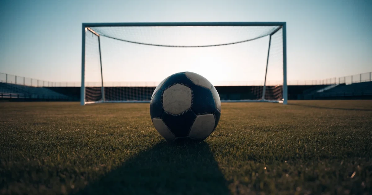 Ein Fußballtor mit Netz im Gegenlicht auf einem leeren Platz am Abend, davor ein Ball auf dem Rasen.