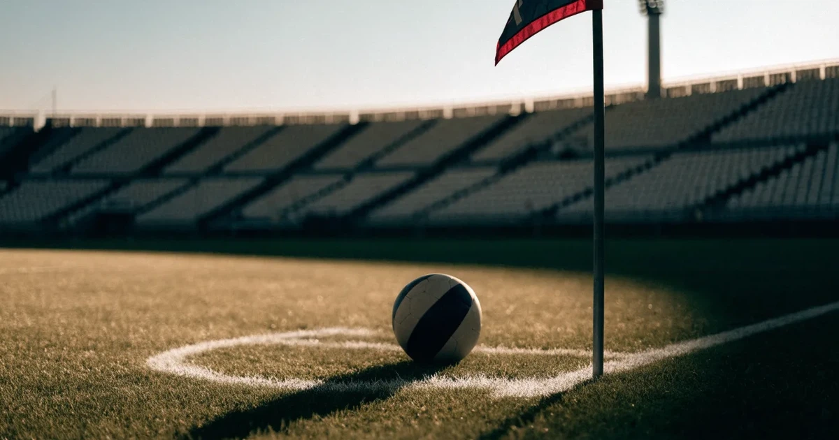 Ein Fußball liegt am Viertelkreis an der Eckfahne eines Rasenplatzes, im Hintergrund leere Tribünen in warmem Abendlicht.