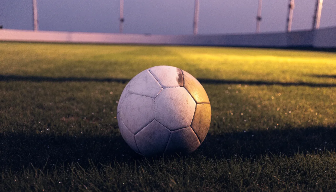 Ein Fußball liegt auf einem Rasenplatz in der Dämmerung, im Hintergrund ein ferner Stadionrand unter Flutlicht.